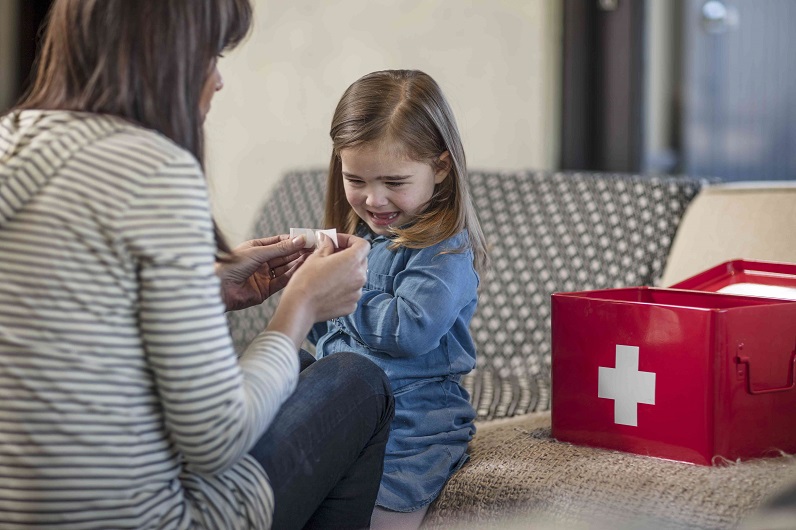picture of a woman putting a bandage on a little girl  who is sitting on a sofa  beside a box first aid kit 