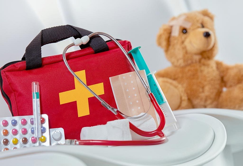 picture of first aid kit essential content beside a bag on a chair with a teddy bear in the background